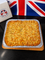 A baked square dish with a textured golden-brown top, presented in a silver foil tray on a wooden board. A white box with a Union Jack logo is visible on the left, and a Union Jack flag sits in the background on a dark surface.