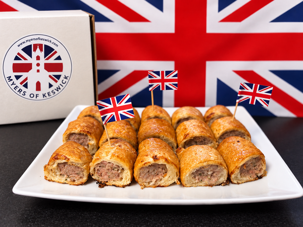 A plate of golden-brown baked sausage rolls with meat filling, each topped with a small Union Jack flag pick, set in front of a large Union Jack flag and next to a branded box reading Myers of Keswick.
