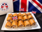 A white rectangular plate filled with golden-brown baked pastries, each topped with a small Union Jack toothpick flag. A branded box reading Myers of Keswick is visible at left, and large Union Jack flags sit in the background on a dark surface.