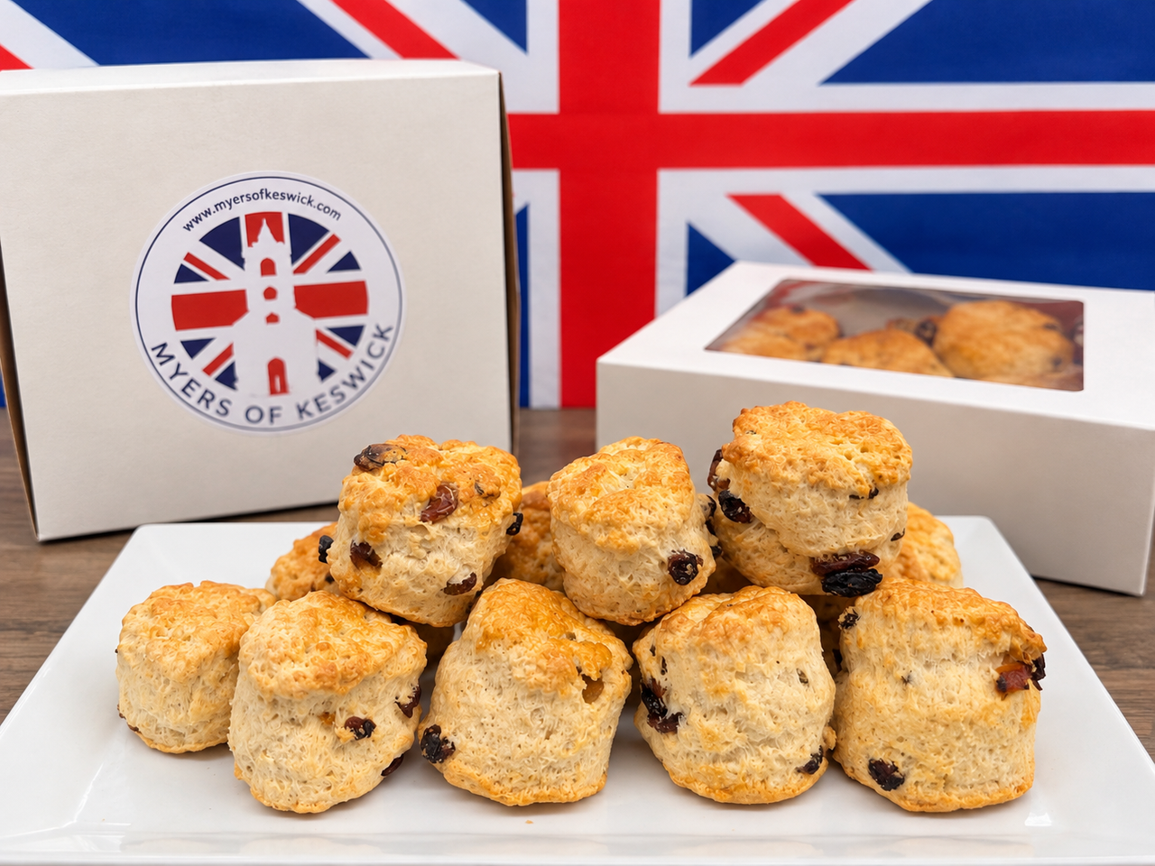A pile of golden-brown scones with raisins on a white plate, with two white product boxes and a Union Jack flag in the background. The front box shows the Myers of Keswick logo.