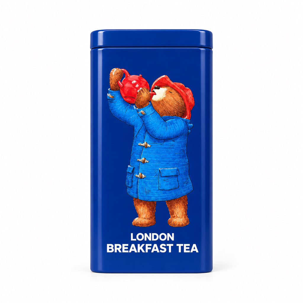 A blue cylindrical tea tin featuring Paddington Bear in a blue duffel coat and red hat, with the text 'LONDON BREAKFAST TEA' on the front, standing on a stone countertop in front of a wooden door.