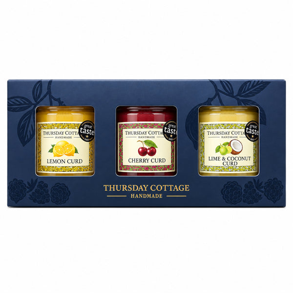 A boxed set of three jars of spread from Thursday Cottage Hampstead, presented in a dark charcoal gift box with leaf illustrations. The set includes Lemon Curd, Chilli Curd, and Lime & Coconut Curd, displayed on a stone ledge against a blurred wooden background.