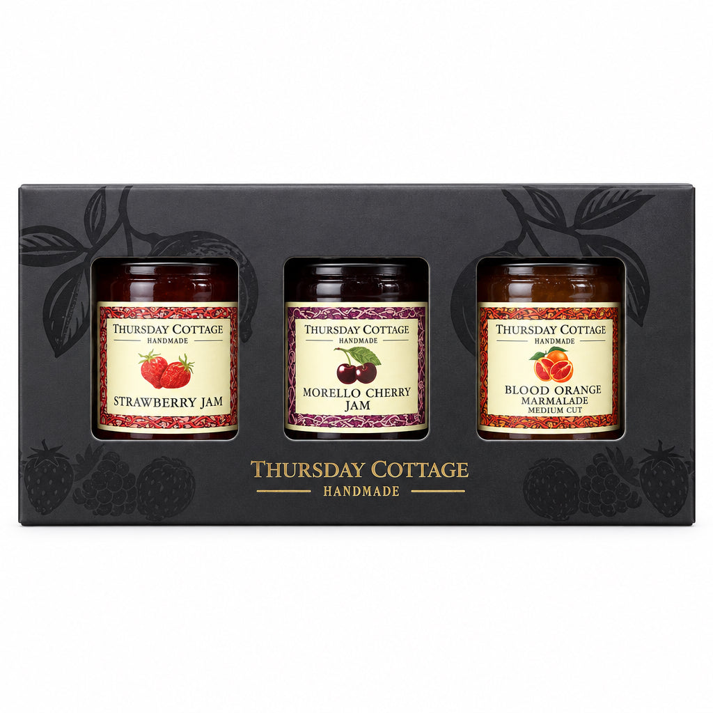 A boxed set of three jars of Thursday Cottage Handmade jam, including Strawberry Jam, Morello Cherry Jam, and Blood Orange Jam, presented in a black gift box on a stone surface with a wooden background.