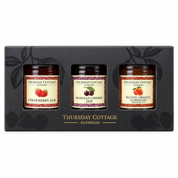 A boxed set of three jars of Thursday Cottage Handmade jam, including Strawberry Jam, Morello Cherry Jam, and Blood Orange Jam, presented in a black gift box on a stone surface with a wooden background.