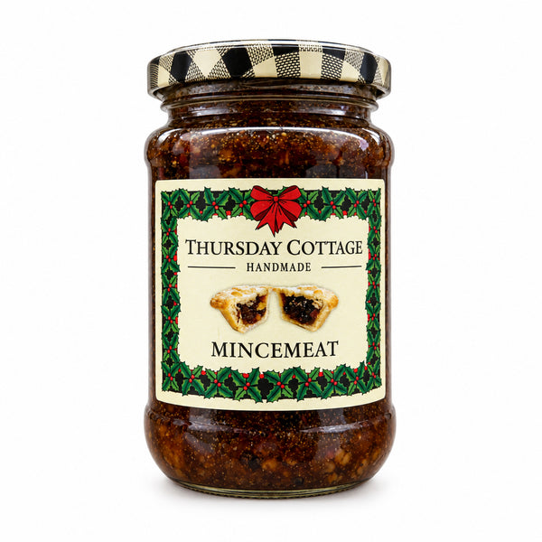 A glass jar of Thursday Cottage Handmade Mincemeat on a white background. The jar contains dark brown mince meat and has a cream-colored label with a red bow and green holly border, topped with a black and white checkered lid.